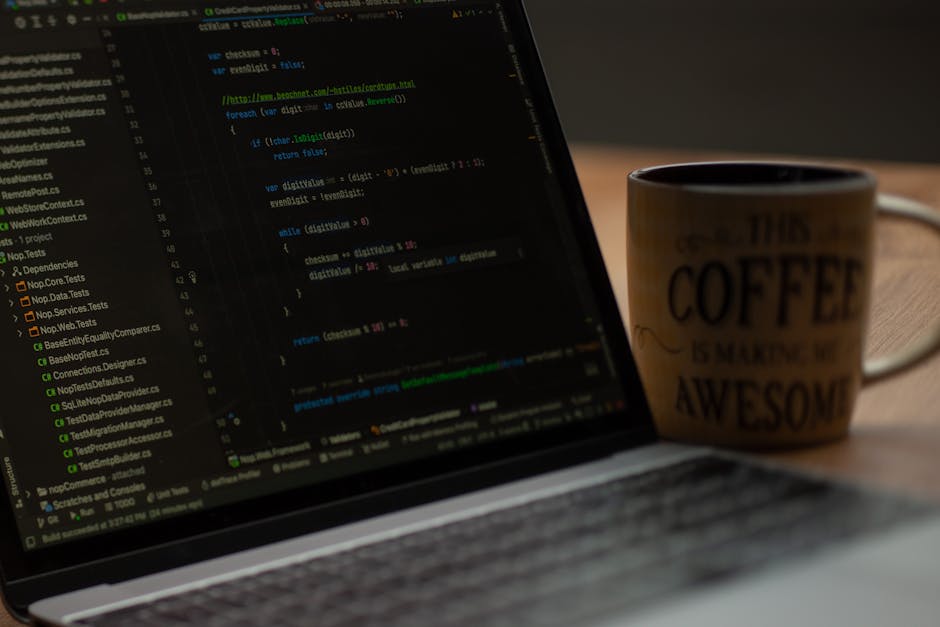 Close-up view of a laptop screen displaying lines of programming code on a dark background, situated on a wooden desk. To the right of the laptop, there is a ceramic coffee mug with black text that re