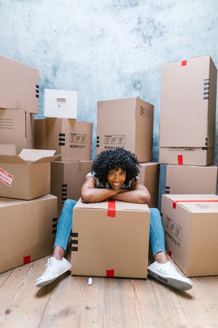 A young woman with curly black hair, wearing a white t-shirt, blue jeans, and white sneakers, is sitting on the wooden floor inside a room surrounded by numerous cardboard boxes of various sizes, some