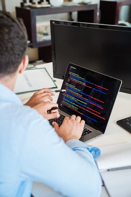 A person is working on a laptop placed on a desk, with hands visible typing on the keyboard. The laptop screen displays lines of code in a dark-themed development environment, including various syntax
