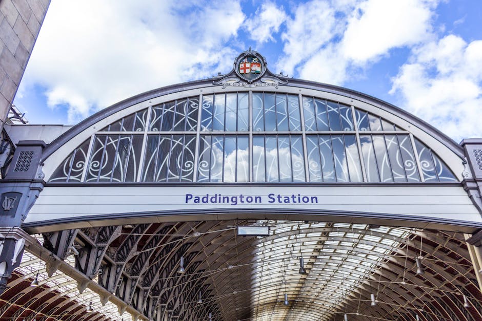 Interior view of a train station with a high, curved glass and metal roof structure allowing natural light to illuminate the platform area. On the platform, there is a yellow and black train approachi